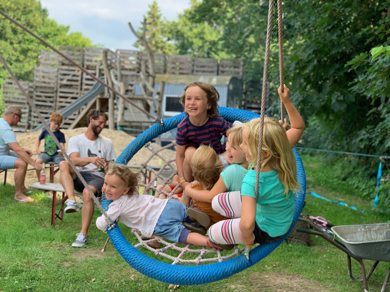 Lachende Kinder schaukeln gemeinsam in einer runden Nestschaukel auf dem Spielplatz, im Hintergrund ein Kletter-Spielturm und Erwachsene auf einer Bank