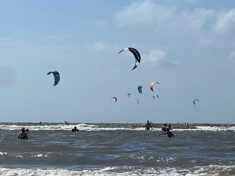 Kitesurfer mit bunten Schirmen über den Wellen der Nordsee, im Vordergrund Badegäste im Wasser