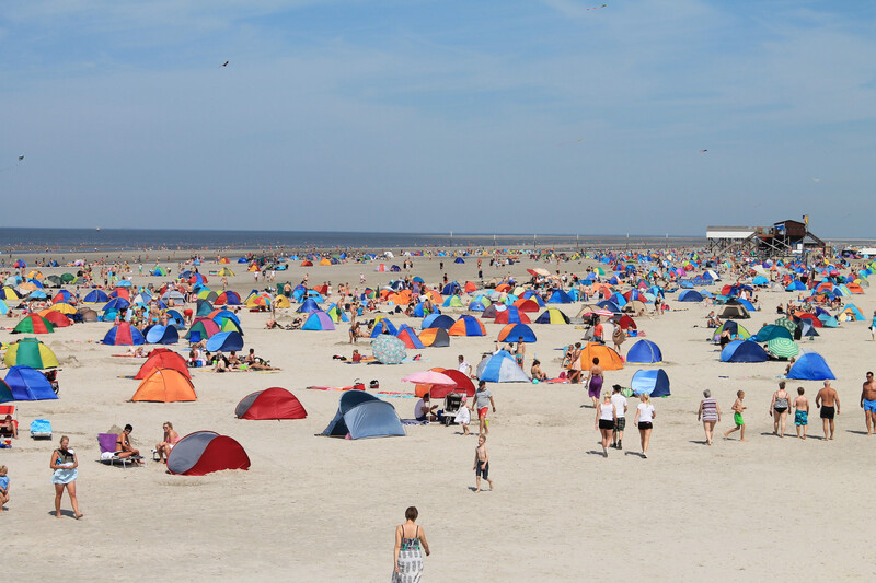 Belebter Sandstrand mit vielen bunten Strandmuscheln, Badegästen und einem Pfahlbau im Hintergrund