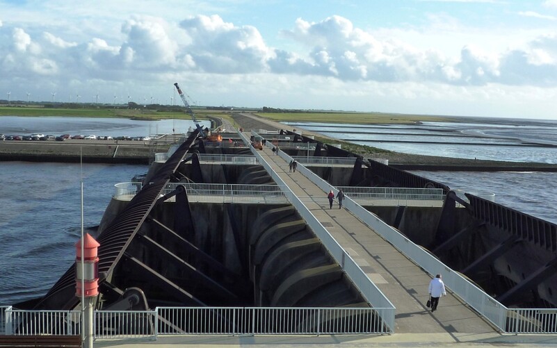 Das Eidersperrwerk an der Nordseeküste mit begehbaren Stegen über den Sielöffnungen, Spaziergängern und Windrädern im Hintergrund