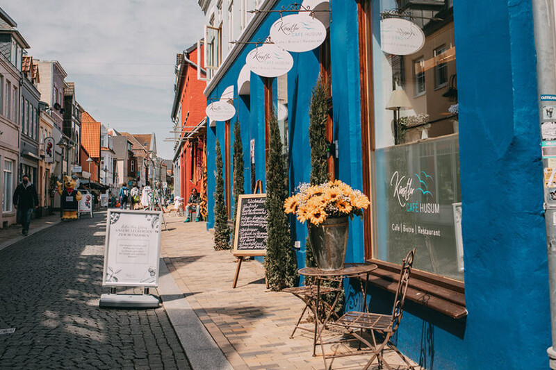 Das blaue Künstler Café Husum in einer gepflasterten Altstadtgasse, mit Sonnenblumen, Bistrotisch und Kreidetafeln vor der Fassade