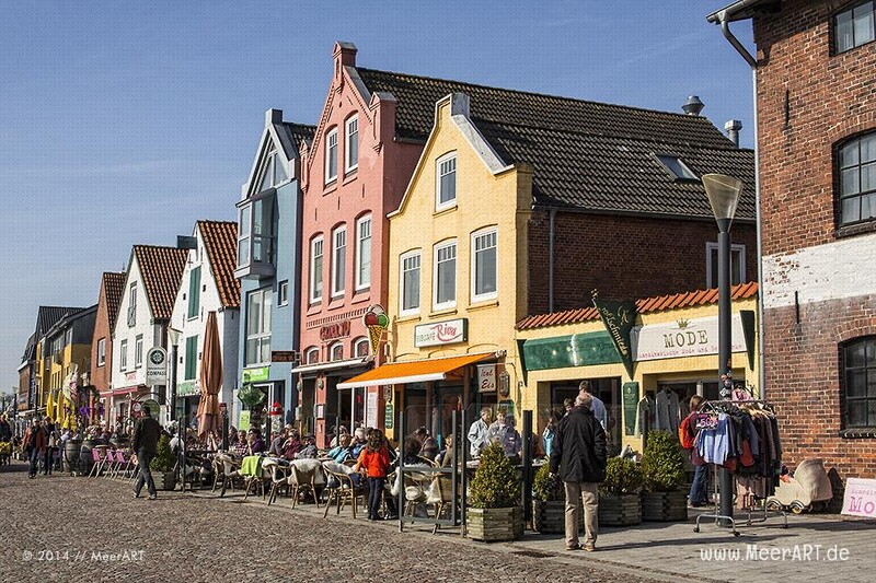 Belebte Hafenpromenade in Husum mit bunten Giebelhäusern, Straßencafés und Geschäften an einem sonnigen Tag