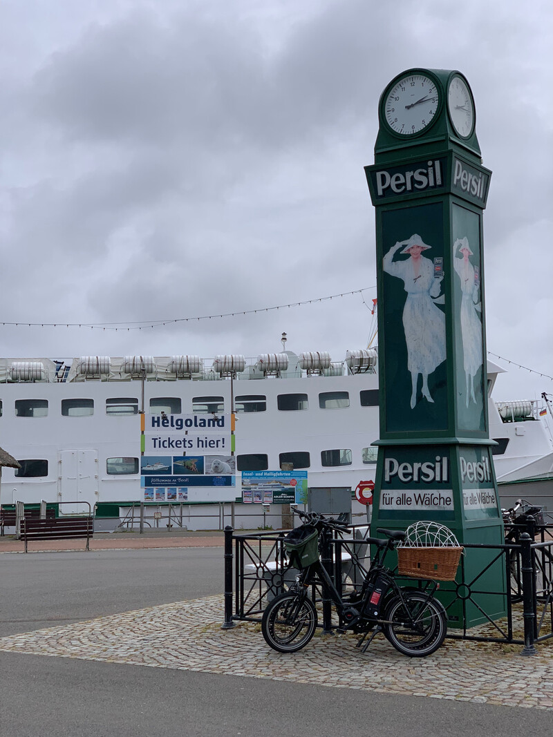 Historische grüne Persil-Uhr am Hafen mit angelehnten Fahrrädern, im Hintergrund ein Fährschiff mit „Helgoland Tickets