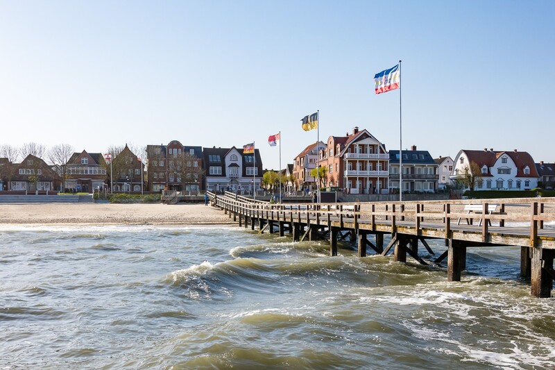 Hölzerne Seebrücke mit wehenden Flaggen führt vom Sandstrand zur Häuserzeile eines Nordsee-Ortes auf der Insel Föhr