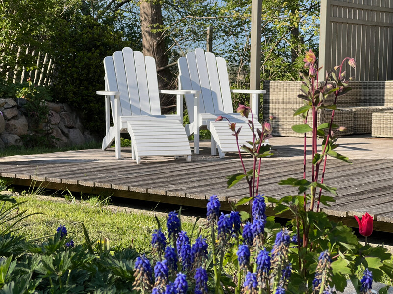 Zwei weiße Holzliegen mit Fußstützen auf einer Holzterrasse, im Vordergrund blühende Traubenhyazinthen und eine Tulpe im Garten