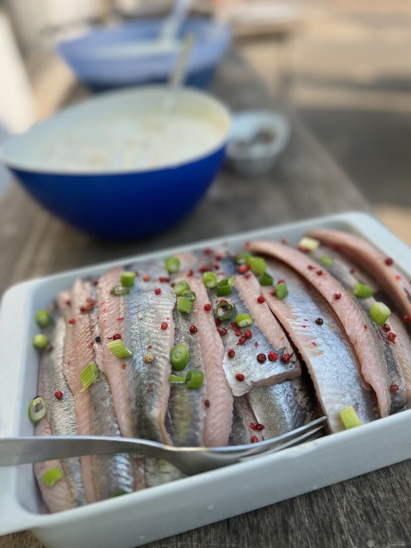 Fischfilets in Schale, silbrige Haut, geschnittene Frühlingszwiebeln, rote Pfefferkörner, Gabel im Vordergrund, Schüssel mit Sauce unscharf im Hintergrund