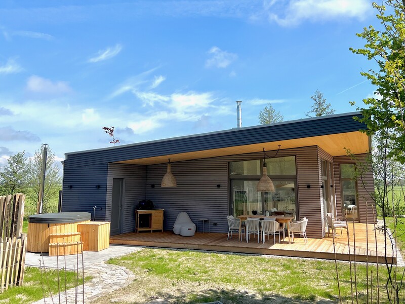 Modernes Holzhaus, Flachdach, große Fensterfront, überdachte Terrasse, Esstisch mit Stühlen, Hängelampen, Hot Tub im Garten, Holzdeck, Rasen, blauer Himmel