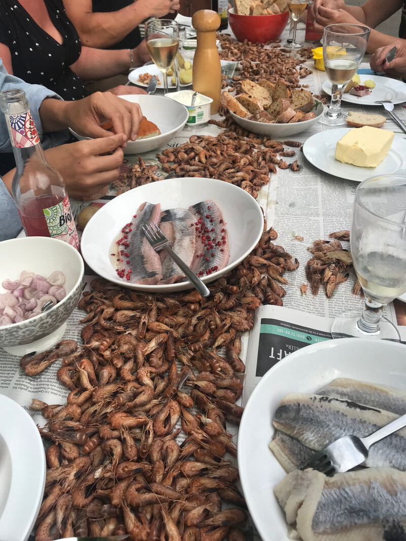 Esstisch im Freien, Zeitung als Unterlage, viele Garnelen, Schalenreste, Schüsseln mit Fischfilets und Zwiebeln, Brot, Butter, Gläser mit Weißwein, mehrere Personen beim Essen