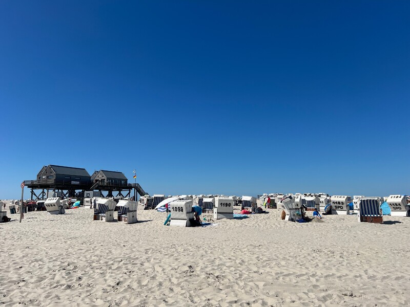 Sandstrand, zahlreiche Strandkörbe mit Nummern, Holzgebäude auf Stelzen mit WC-Schild, wenige Menschen, klarer blauer Himmel