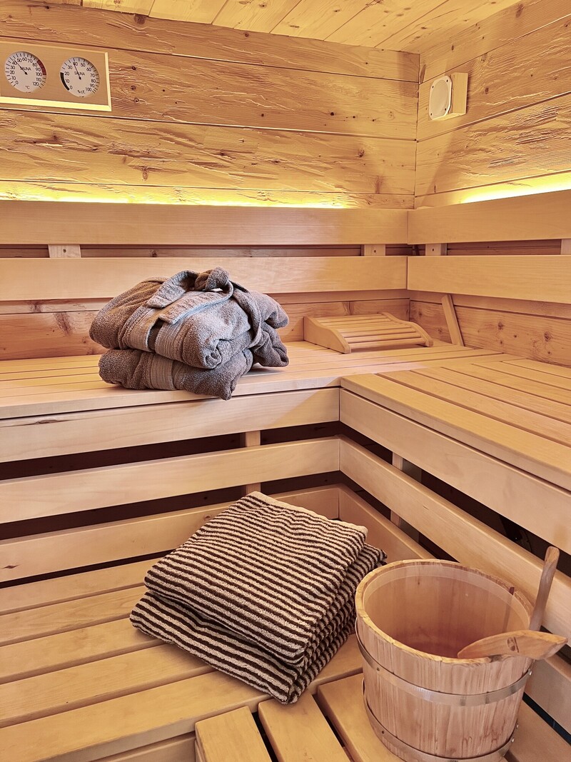 Sauna-Innenraum, helle Holzverkleidung, zwei Sitzbänke, gefaltete Handtücher, Holzeimer mit Kelle, indirekte Beleuchtung, Thermometer an der Wand