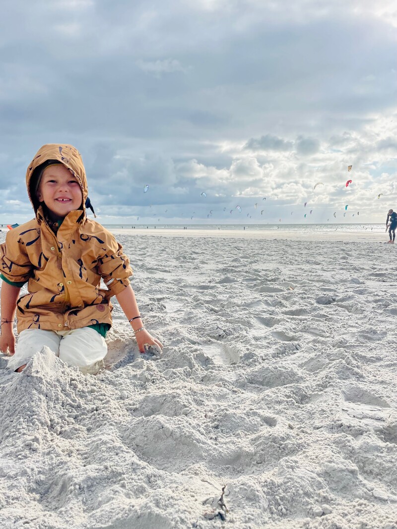 Kind am Strand, sitzt im Sand, braune Kapuzenjacke, heller Sandstrand, Meer im Hintergrund, viele Kiteschirme am Himmel, bewölkter Himmel