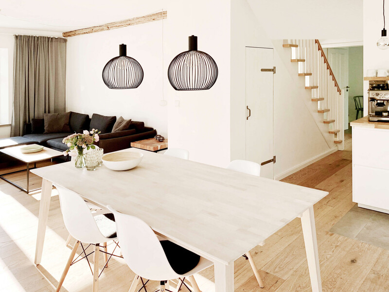 Helles Wohn-Esszimmer, großer Holztisch, weiße Stühle, schwarze Draht-Hängelampen, graues Sofa, Couchtisch, Blumenstrauß, Holzfußboden, offene Treppe, Küche mit Kaffeemaschine