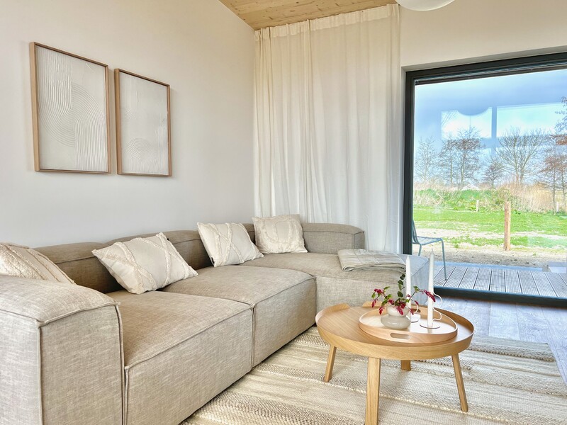 Wohnzimmer, hell, L-Sofa mit Kissen, runder Holztisch, Kerzenständer, kleine Vase mit Blumen, Teppich, große Glas-Schiebetür, Blick ins Grüne, Vorhänge, Holzdecke