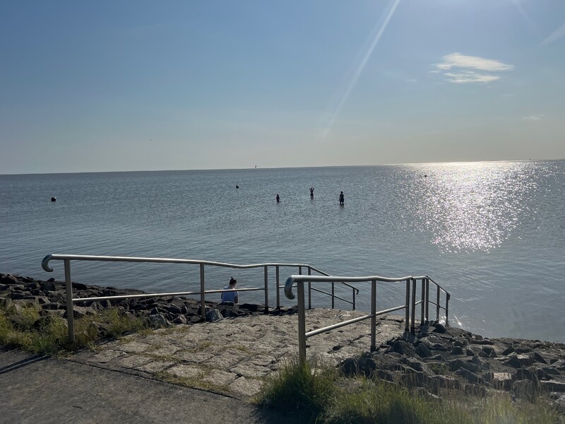 Küstenzugang, Steintreppe mit Geländer, ruhiges Meer, drei Personen im Wasser, Sonnenreflexion, weiter Horizont, klarer Himmel