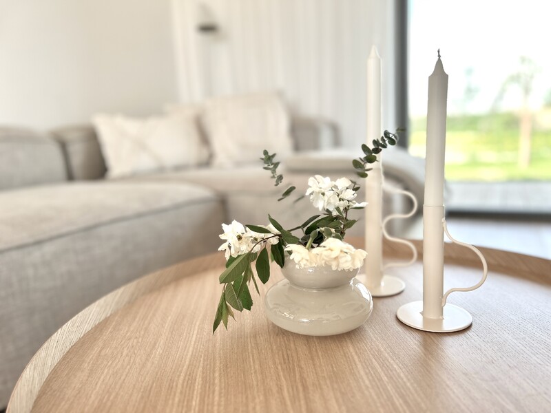Wohnzimmer, runder Holztisch, weiße Kerzenständer, Vase mit weißen Blumen, Sofa im Hintergrund, große Fensterfront, helle, minimalistische Einrichtung