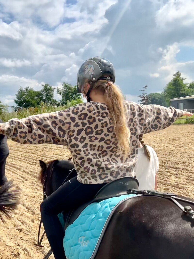 Kind reitet Pony, Arme ausgestreckt, Reithelm, Sandplatz, Leopardmuster-Pullover, Pferderücken im Vordergrund, Himmel mit Wolken
