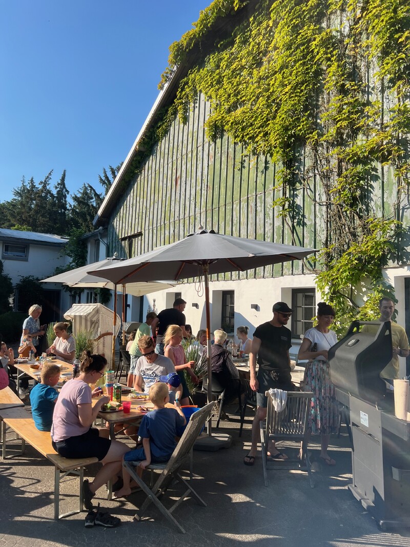 Außenbereich mit langen Holztischen, viele Menschen, Familien mit Kindern, Sonnenschirme, Grillstation, großes Gebäude mit begrünter Holzfassade, sonniger Himmel