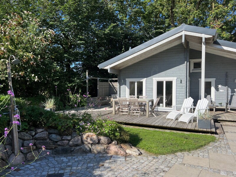 Modernes Holzhaus, graue Fassade, große Terrasse, Esstisch mit Stühlen, zwei Liegestühle, Garten mit Blumen, Bäume im Hintergrund, gepflasterter Weg, Außendusche