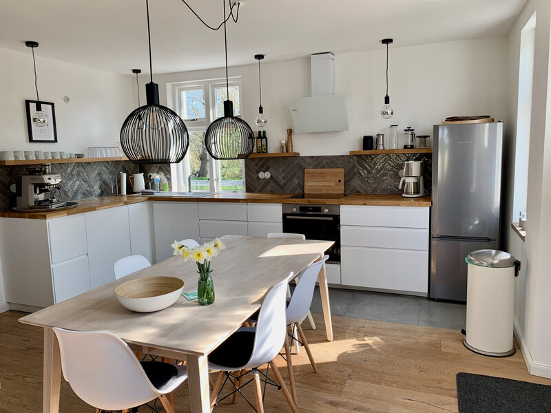 Helle, moderne Küche mit weißer Einrichtung, Holzarbeitplatte und Esstisch; mehrere Hängelampen, Kühlschrank und große Fenster sorgen für ein freundliches Ambiente