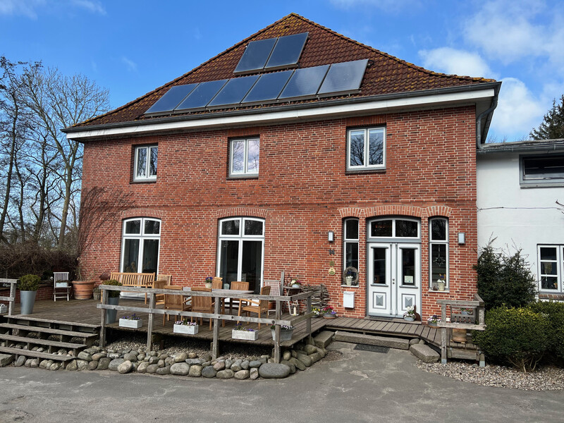 Zweistöckiges Backsteinhaus mit rotem Ziegeldach und mehreren Solarpaneelen; vor dem Haus eine Holzterrasse mit Sitzmöbeln und Pflanzen