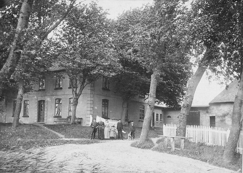 Schwarzweißfotografie eines Hauses mit mehreren Personen, die vor einem weißen Zaun stehen; umgeben von Bäumen und einem Hofweg