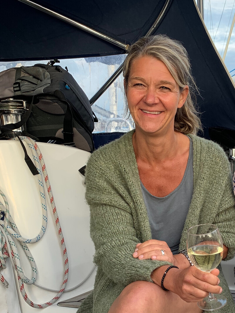 Eine lächelnde Frau sitzt auf einem Boot, hält ein Glas Weißwein in der Hand und blickt in die Kamera