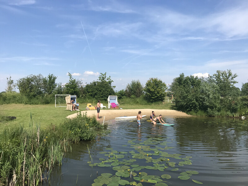 Mehrere Menschen stehen und spielen an einem kleinen Badeteich; einige sind im Wasser mit Surfbrettern, im Vordergrund schwimmen Seerosen, im Hintergrund stehen Strandkörbe und Bäume