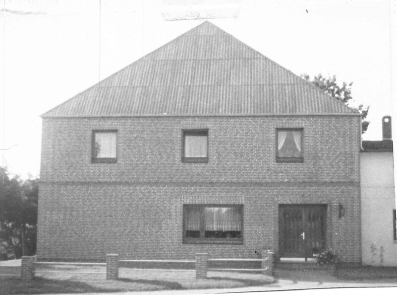 Schwarzweißfoto eines zweistöckigen Backsteinhauses mit Walmdach; schlichte Fassade mit wenigen Fenstern, kleiner Vorgarten und niedriger Zaun davor