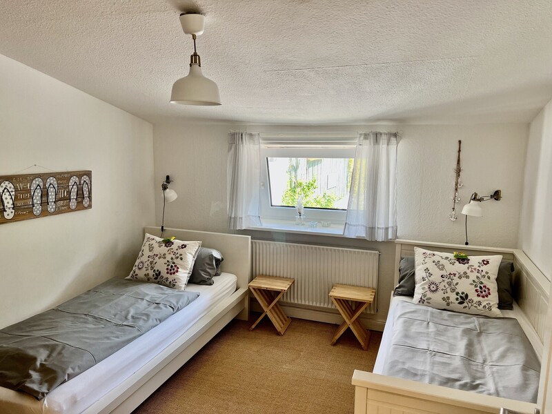 Schlafzimmer mit zwei Einzelbetten links und rechts, grauer Bettwäsche und geblümten Kissen, Leselampen und Fenster mit Blick ins Grüne