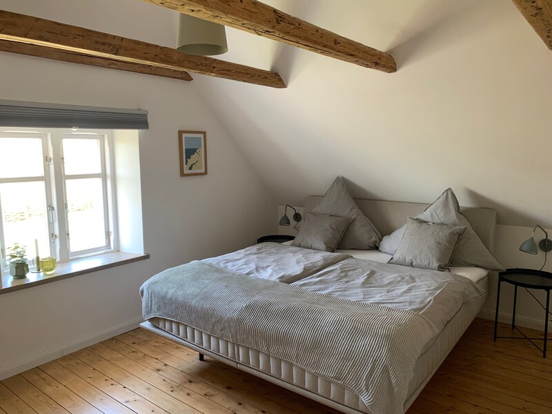 Helles Schlafzimmer unter der Dachschräge mit Boxspring-Doppelbett, rustikalen Holzdeckenbalken und Dielenboden; Fenster mit Tageslicht