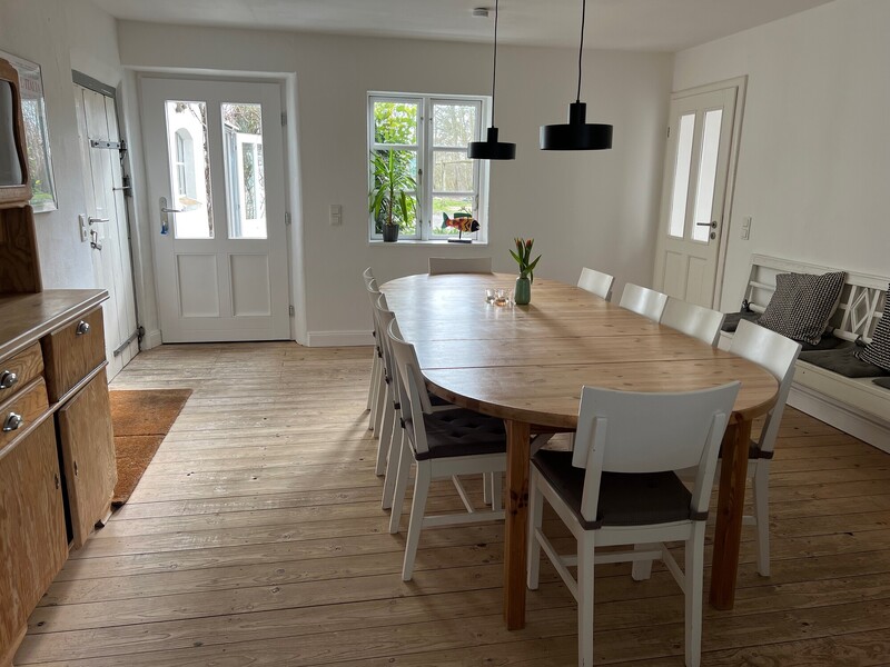 Heller Essbereich mit großem ovalem Holztisch und weißen Stühlen, Sitzbank, Holzbuffet und Dielenboden; Sprossenfenster und Tür mit Blick nach draußen