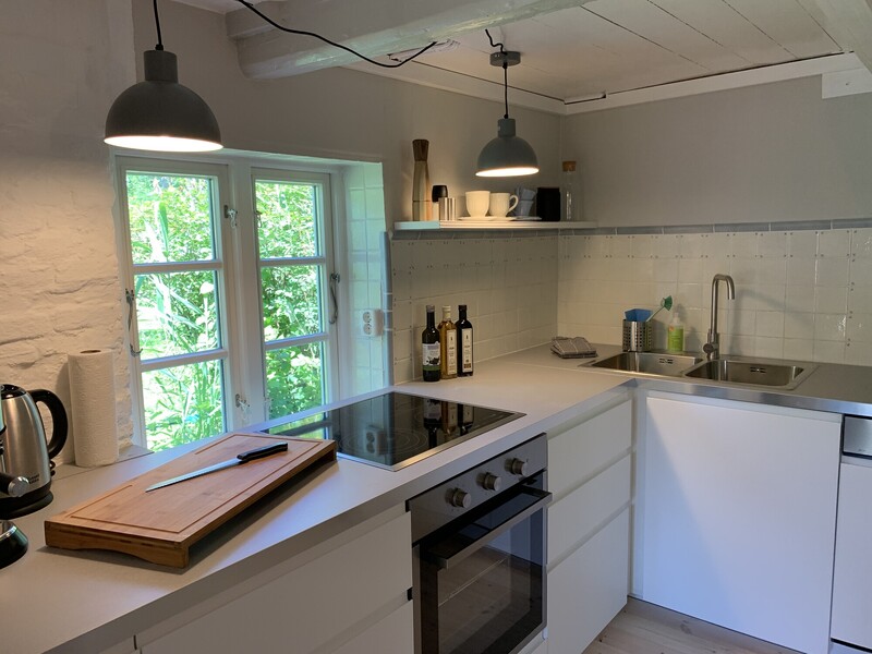 Helle Küche mit weißer Einbauküche, Ceranfeld, Backofen und Spüle; Holzschneidebrett auf der Arbeitsplatte, Sprossenfenster mit Blick ins Grüne und weiß gestrichene Deckenbalken