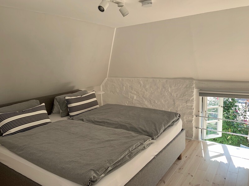 Schlafzimmer unter der Dachschräge mit Doppelbett, grauer Bettwäsche und gestreiften Kissen; weiß gestrichene Natursteinwand und Fenster mit Blick ins Grüne