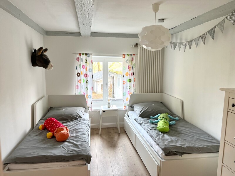 Kinderzimmer mit zwei Einzelbetten, Kuscheltieren, Wimpelkette und weiß gestrichenen Deckenbalken; Fenster mit Blick auf ein Nachbarhaus