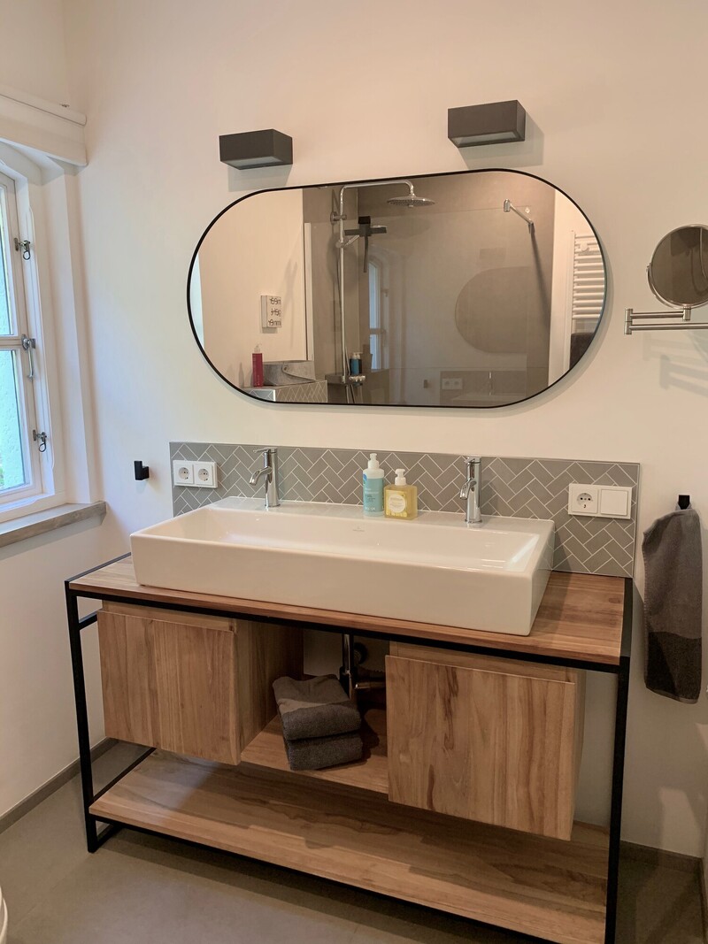 Modernes Badezimmer mit breitem Doppelwaschbecken auf Holz-Metall-Waschtisch, ovalem Spiegel und grauem Fischgrätfliesenspiegel; im Spiegel die Walk-in-Dusche