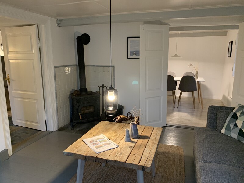Wohnzimmer mit gusseisernem Kaminofen, grauem Sofa und rustikalem Holz-Couchtisch; Blick durch die offene Tür in den Essbereich