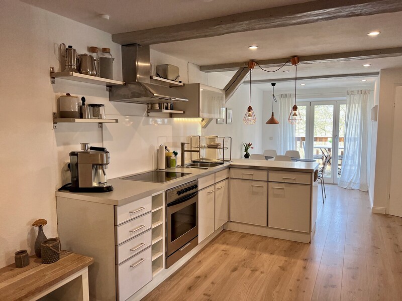 Offene L-Küche in Beige mit Ceranfeld, Backofen, Edelstahldunstabzug und offenen Regalen; sichtbarer Deckenbalken und Essbereich mit Terrassentür im Hintergrund