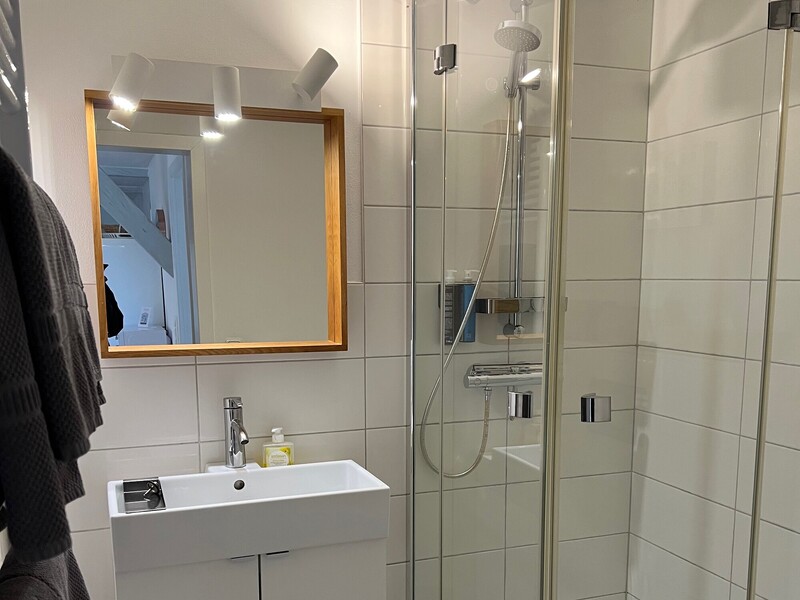 Weiß gefliestes Badezimmer mit Dusche hinter Glastür, Waschbecken und holzgerahmtem Spiegel