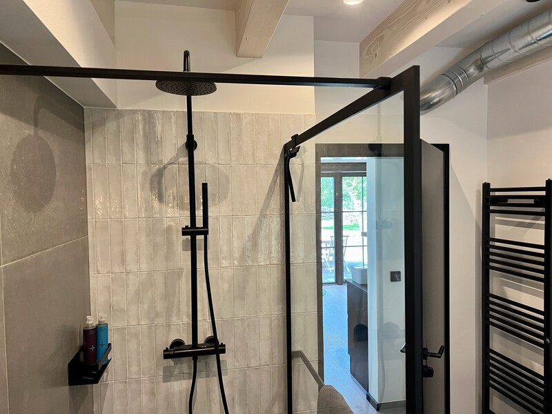 Bodengleiche Walk-in-Dusche mit schwarzer Regendusche und Glaswand, hell gefliest; sichtbare Deckenbalken und Blick in den angrenzenden Waschbereich