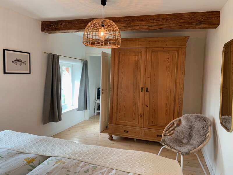 Landhaus-Schlafzimmer mit Doppelbett, antikem Holzkleiderschrank, Rattansessel mit Fell und rustikalen Holzdeckenbalken; Fenster mit Tageslicht