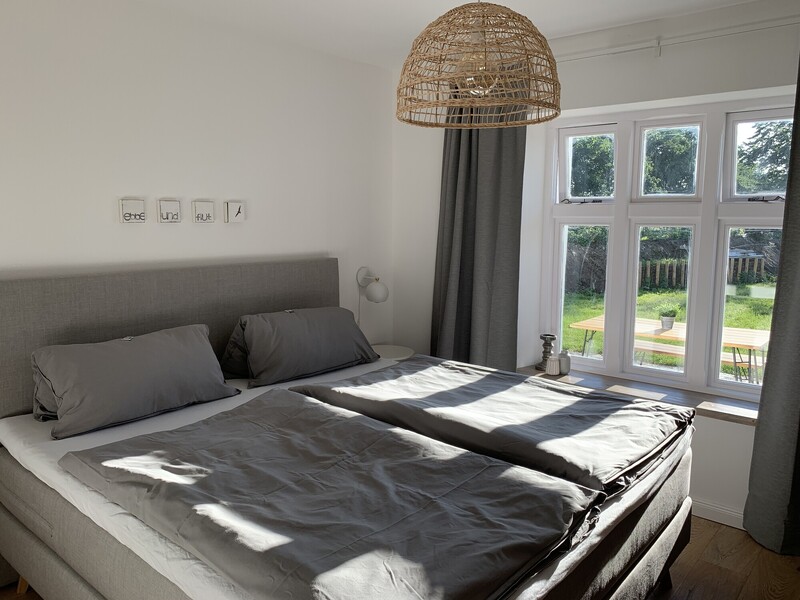 Helles Schlafzimmer mit Doppelbett, grauer Bettwäsche, Korblampe und Sprossenfenster mit Blick auf Garten und Terrasse