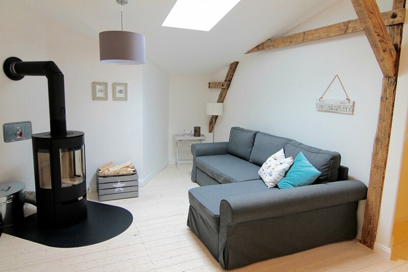 Wohnbereich einer Ferienunterkunft mit Sofa, Kaminofen, Holzbalken und Dachfenster am Warfthof Wollatz