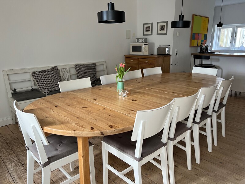 Großer ovaler Holzesstisch mit weißen Stühlen und Sitzbank mit Kissen, im Hintergrund Buffet und offene Küche mit Dielenboden