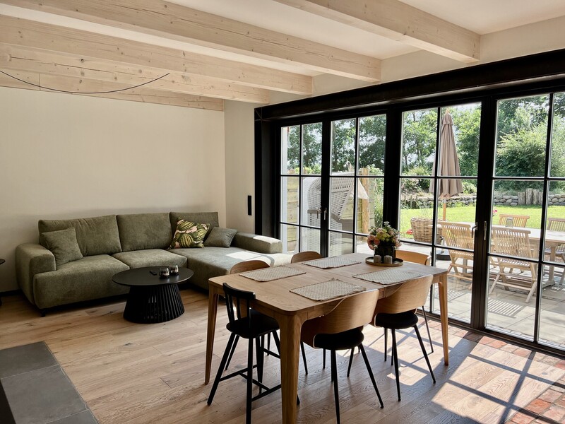 Heller Wohn-Essbereich mit grünem Ecksofa, Holzesstisch und Panoramafenster mit Blick auf Terrasse, Strandkorb und Garten; helle Holzdeckenbalken