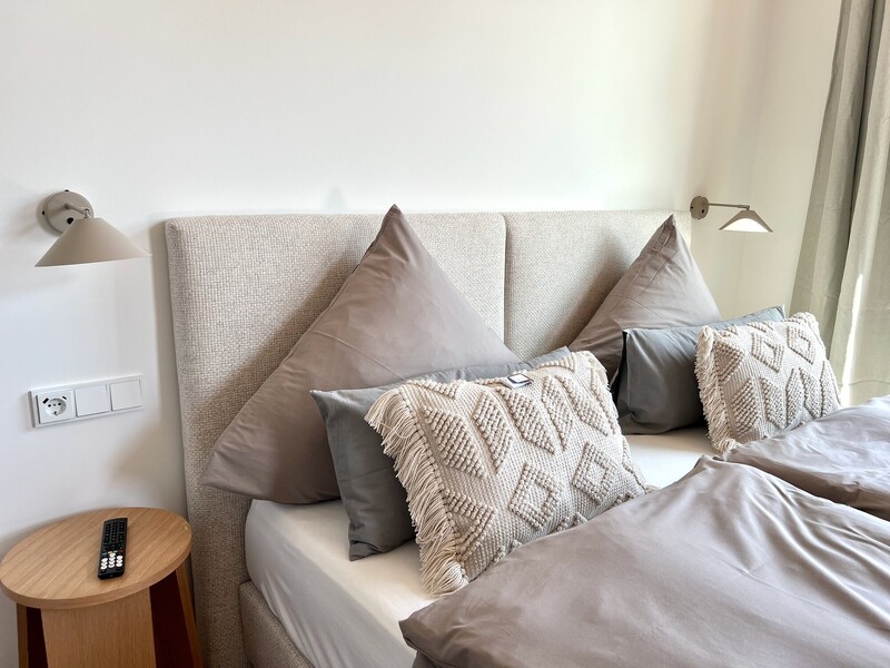 Doppelbett mit gepolstertem Kopfteil, grauer Bettwäsche und dekorativen Kissen, Nachttisch und Leselampen
