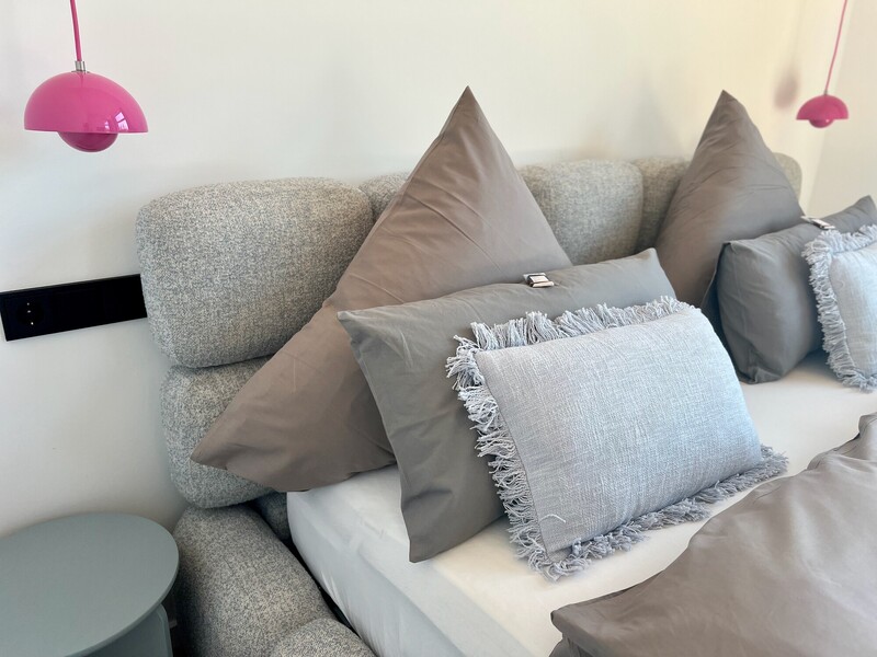 Bett-Detail im Haus NordseeGut mit grauen Kissen, gepolstertem Kopfteil und pinken Pendelleuchten