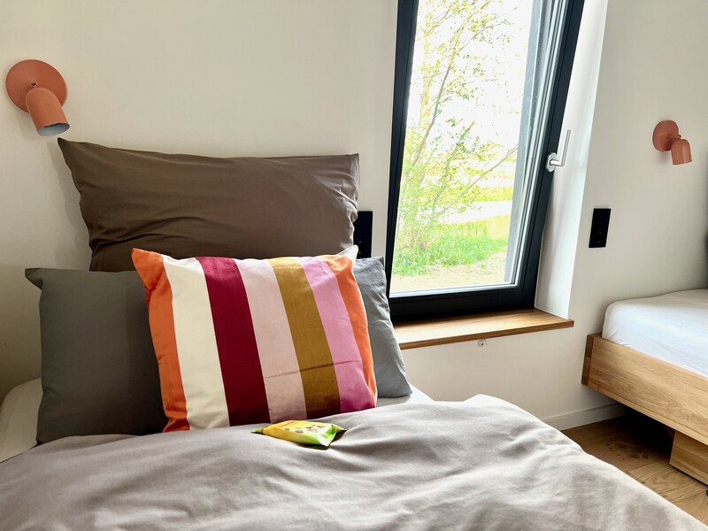 Bett-Detail im Haus NordseeGut mit gestreiftem Kissen, Wandleuchten und Fenster mit Blick ins Grüne