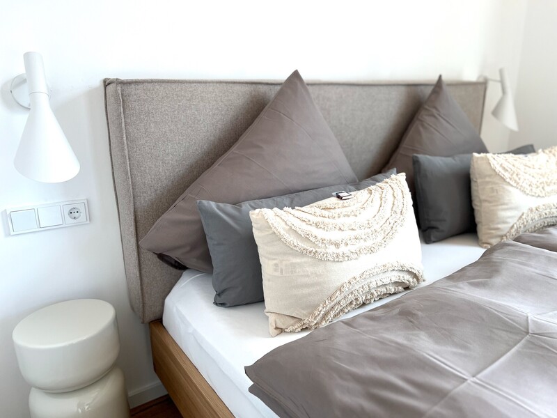 Bett-Detail vom Haus NordseeLuft mit grauen Kissen, gepolstertem Kopfteil und weißer Wandleuchte