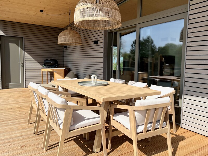 Überdachte Terrasse vom Haus NordseeZeit mit Holz-Esstisch, Stühlen, Rattanleuchten und Grill in der Sonne