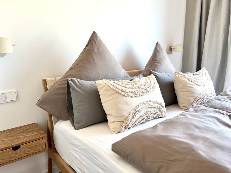 Bett-Detail im Haus NordseeZeit mit Kissen, geflochtenem Holz-Kopfteil, Nachttisch und Wandleuchte